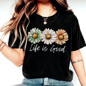 May include: Black t-shirt featuring three daisy flowers in pastel colors: green, white, and orange. The words "Life is Good" are written in white script below the flowers. The shirt is a casual, everyday wear item.