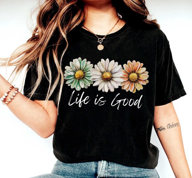 May include: Black t-shirt featuring three daisy flowers in pastel colors: green, white, and orange. The words "Life is Good" are written in white script below the flowers. The shirt is a casual, everyday wear item.