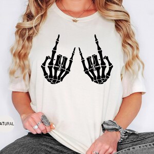 May include: A cream-colored short-sleeve t-shirt featuring a black graphic print of two skeleton hands making the rock and roll hand sign. The shirt has a crew neckline and the word NATURAL is printed in small black text on the lower left side.