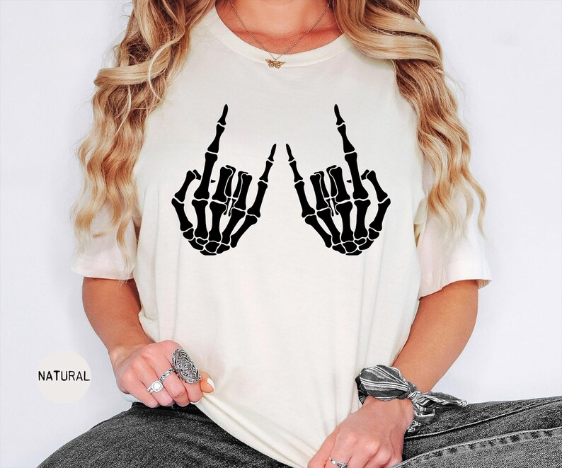 May include: A cream-colored short-sleeve t-shirt featuring a black graphic print of two skeleton hands making the rock and roll hand sign. The shirt has a crew neckline and the word NATURAL is printed in small black text on the lower left side.