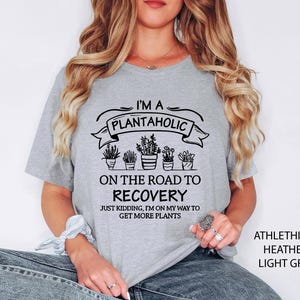 Op de afbeelding: Lichtgrijs t-shirt met de tekst "I'M A PLANTAHOLIC ON THE ROAD TO RECOVERY" en een afbeelding van potplanten. Het shirt bevat ook de zin "JUST KIDDING, I'M ON MY WAY TO GET MORE PLANTS".