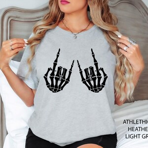 May include: A light grey t-shirt featuring a black graphic print of two skeleton hands making the rock and roll hand sign. The text ATHLETHICH HEATHER LIGHT GREY is printed in the bottom right corner of the image.