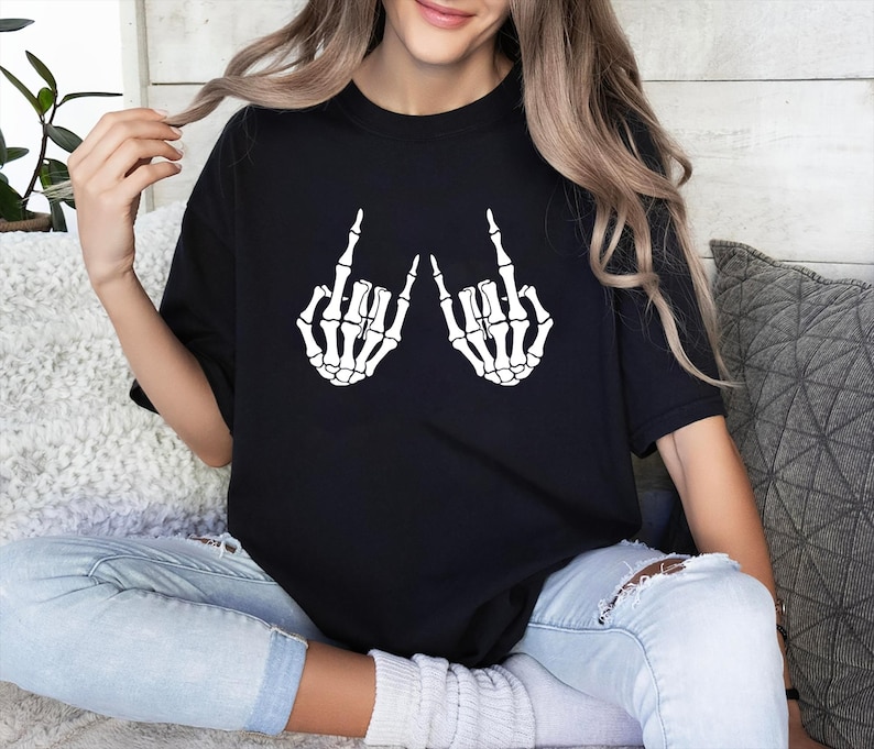 May include: A black short-sleeved t-shirt featuring a white graphic print of two skeletal hands making the rock and roll hand gesture. The design is centered on the chest of the shirt, which has a crew neckline and a relaxed, loose-fitting cut.