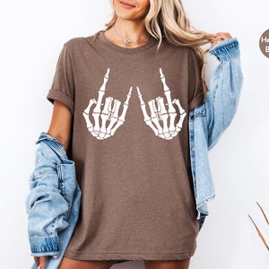 May include: A heather brown short-sleeve t-shirt featuring a white graphic print of two skeleton hands making a rock and roll hand gesture. The shirt has a crew neckline and a relaxed fit, with a small circular badge in the corner labeled Heather Brown.