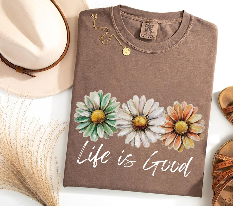 May include: A brown t-shirt with a daisy floral design in shades of green, white, and orange. The text "Life is Good" is printed in white script below the flowers. A gold necklace and a hat are next to the shirt.