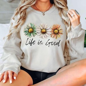 K&ouml;nnte beinhalten: Cremefarbenes Sweatshirt mit drei G&auml;nsebl&uuml;mchen in Gr&uuml;n, Wei&szlig; und Orange mit gelben Zentren. Der Schriftzug "Life is Good" steht in schwarzer Schrift darunter. Das Sweatshirt hat lange &Auml;rmel.