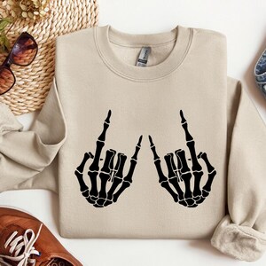 May include: A beige crewneck sweatshirt featuring a black graphic print of two skeleton hands making the rock and roll hand sign. The sweatshirt has long sleeves, a ribbed collar, and a standard fit, showcasing a clear contrast between the fabric.