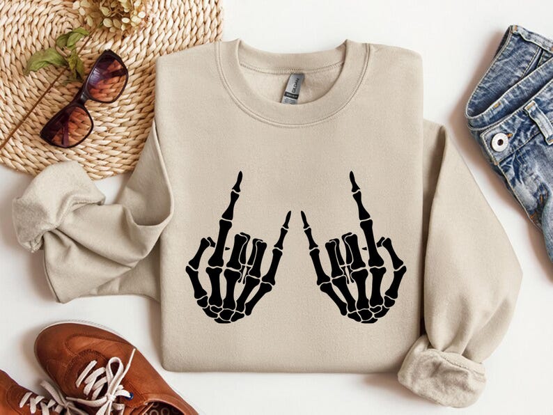 May include: A beige crewneck sweatshirt featuring a black graphic print of two skeleton hands making the rock and roll hand sign. The sweatshirt has long sleeves, a ribbed collar, and a standard fit, showcasing a clear contrast between the fabric.