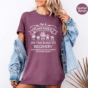 Op de afbeelding: Bordeaux heather t-shirt met witte tekst: "I'M A PLANTAHOLIC ON THE ROAD TO RECOVERY JUST KIDDING, I'M ON MY WAY TO GET MORE PLANTS". Het shirt heeft een afbeelding van potplanten. Een spijkerjasje wordt over het shirt gedragen.