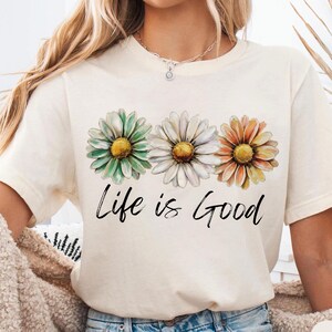 K&ouml;nnte beinhalten: Cremefarbenes T-Shirt mit drei G&auml;nsebl&uuml;mchen in Gr&uuml;n, Wei&szlig; und Orange mit gelben Zentren. Der Schriftzug "Life is Good" steht in schwarzer Schrift darunter. Das Shirt hat kurze &Auml;rmel.