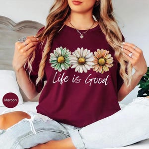 K&ouml;nnte beinhalten: Burgunderfarbenes T-Shirt mit drei G&auml;nsebl&uuml;mchen in Gr&uuml;n, Wei&szlig; und Orange mit gelben Zentren. Die Worte "Life is Good" sind darunter in wei&szlig;er Schrift geschrieben. Das Shirt hat einen Rundhalsausschnitt.