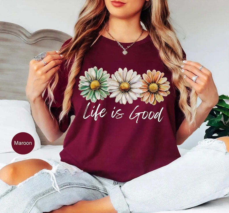 K&ouml;nnte beinhalten: Burgunderfarbenes T-Shirt mit drei G&auml;nsebl&uuml;mchen in Gr&uuml;n, Wei&szlig; und Orange mit gelben Zentren. Die Worte "Life is Good" sind darunter in wei&szlig;er Schrift geschrieben. Das Shirt hat einen Rundhalsausschnitt.