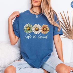 K&ouml;nnte beinhalten: Indigoblaues T-Shirt mit einem Blumendesign aus drei G&auml;nsebl&uuml;mchen in Gr&uuml;n, Wei&szlig; und Orange. Die Worte "Life is Good" sind in einer Schreibschrift unter den Blumen aufgedruckt. Das Shirt ist ein l&auml;ssiger Stil mit kurzen &Auml;rmeln.