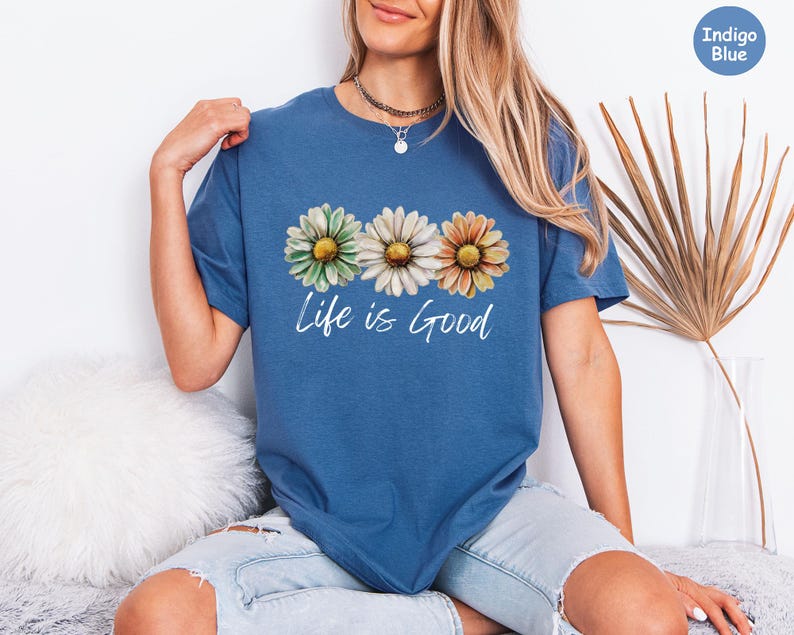 K&ouml;nnte beinhalten: Indigoblaues T-Shirt mit einem Blumendesign aus drei G&auml;nsebl&uuml;mchen in Gr&uuml;n, Wei&szlig; und Orange. Die Worte "Life is Good" sind in einer Schreibschrift unter den Blumen aufgedruckt. Das Shirt ist ein l&auml;ssiger Stil mit kurzen &Auml;rmeln.
