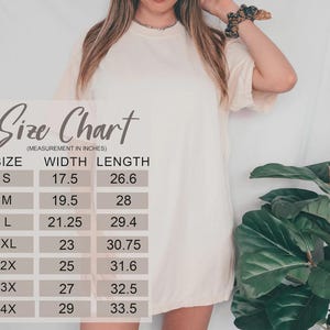 May include: Cream-colored t-shirt with a size chart overlay. The chart lists sizes S through 4X, with width and length measurements in inches. The shirt is worn by a person, with a scrunchie on their wrist.