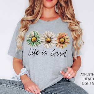 K&ouml;nnte beinhalten: Hellgraues T-Shirt mit drei G&auml;nsebl&uuml;mchen in Gr&uuml;n, Wei&szlig; und Orange und dem Schriftzug "Life is Good" in schwarzer Schrift. Das Shirt besteht aus einem weichen, melierten Material.