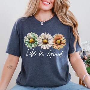 K&ouml;nnte beinhalten: Marineblaues T-Shirt mit drei G&auml;nsebl&uuml;mchen in Gr&uuml;n, Wei&szlig; und Orange mit gelben Zentren. Die Worte "Life is Good" sind in einer Schreibschrift unter den Blumen geschrieben. Das T-Shirt hat kurze &Auml;rmel.