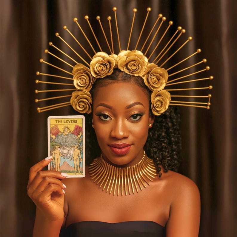 May include: A woman wearing a gold headpiece with rose accents and a matching gold collar. She holds a tarot card labeled "THE LOVERS". The headpiece has radiating gold rods. The woman is wearing a black strapless top.
