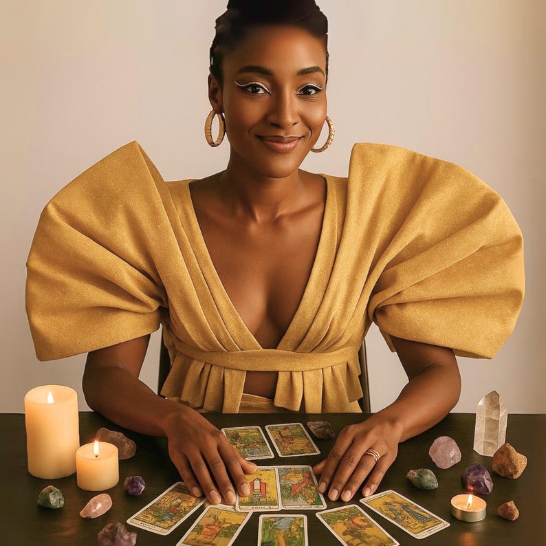 May include: A woman in a mustard-yellow dress with large, structured sleeves sits at a table. Tarot cards are laid out, surrounded by crystals and lit candles. The woman is smiling, wearing gold hoop earrings.