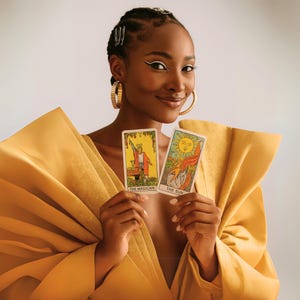 May include: A person holding two tarot cards, "The Magician" and "The Sun." The person is wearing a gold-colored top and large gold hoop earrings. The background is a solid, neutral color.