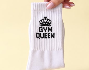 May include: A white ribbed crew sock featuring a black embroidered graphic of a crown positioned above the text "GYM QUEEN" in a bold, sans-serif font. The sock is made of a knit fabric and has a standard length with a reinforced cuff.