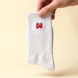 Könnte beinhalten: Eine weiße, gerippte Socke mit einem kleinen, aufgestickten roten Krebsmotiv im oberen Bereich. Die Socke weist eine klassische Strickstruktur auf und besticht durch ein schlichtes, klares Design mit einem detaillierten Krustentier-Motiv.