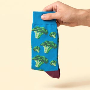 May include: A bright blue crew sock featuring a repeating pattern of green broccoli florets. The sock has a ribbed cuff at the top and a solid dark maroon heel section visible at the bottom, showcasing a vibrant vegetable-themed graphic design.