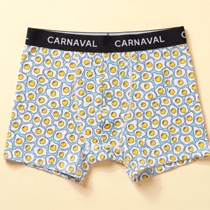 Puede incluir: Calzoncillos tipo bóxer con fondo azul claro y estampado de huevos fritos con yemas amarillas y claras blancas. La cintura elástica negra presenta el texto blanco CARNAVAL repetido en la parte delantera.