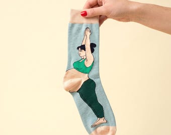 May include: A light blue crew sock featuring a graphic illustration of a person with dark hair wearing a green sports bra and dark green leggings, performing a yoga pose with arms extended upward and a visible baby bump.