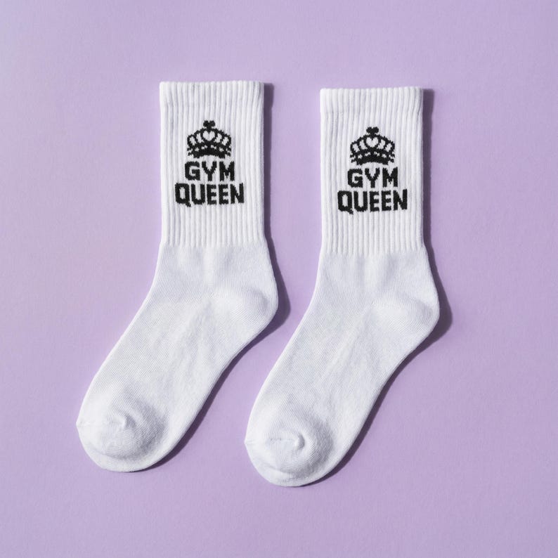 May include: A pair of white crew socks featuring a black graphic of a crown above the text "GYM QUEEN" printed on the ribbed upper section. The socks have a standard knit texture and are displayed side by side against a solid background.