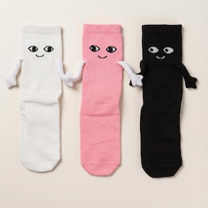 May include: Three crew socks in white, pink, and black, each featuring a cartoon face with two eyes and a small smile. The socks have small fabric arms attached to the sides, with the white and pink socks holding hands, and the pink and black socks holding hands.