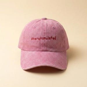 Puede incluir: Gorra de b&eacute;isbol rosa polvoriento con la palabra "overstimulated" bordada en hilo rojo m&aacute;s oscuro en la parte delantera. La gorra tiene una visera curva y un bot&oacute;n en la parte superior.
