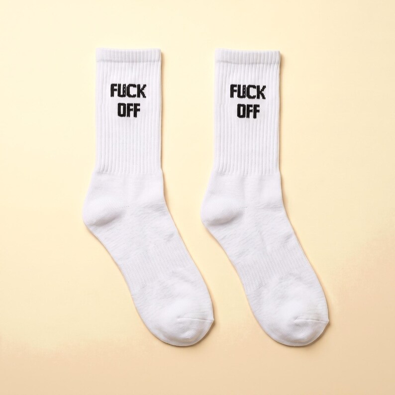 May include: A pair of white crew socks featuring a ribbed texture on the upper section. The text FUCK OFF is printed in bold black capital letters on the front of each sock, positioned vertically on the ribbed cuff area.