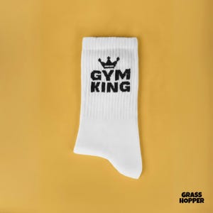 Könnte beinhalten: Weißer Sportsocken mit dem schwarzen Text „GYM KING“ und einer Kronengrafik. Der Socken befindet sich vor einem senfgelben Hintergrund.