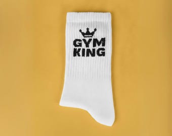 May include: A white crew sock featuring a ribbed texture and a black embroidered graphic on the upper portion. The graphic consists of a crown icon positioned above the bold, capitalized black text that reads GYM KING.