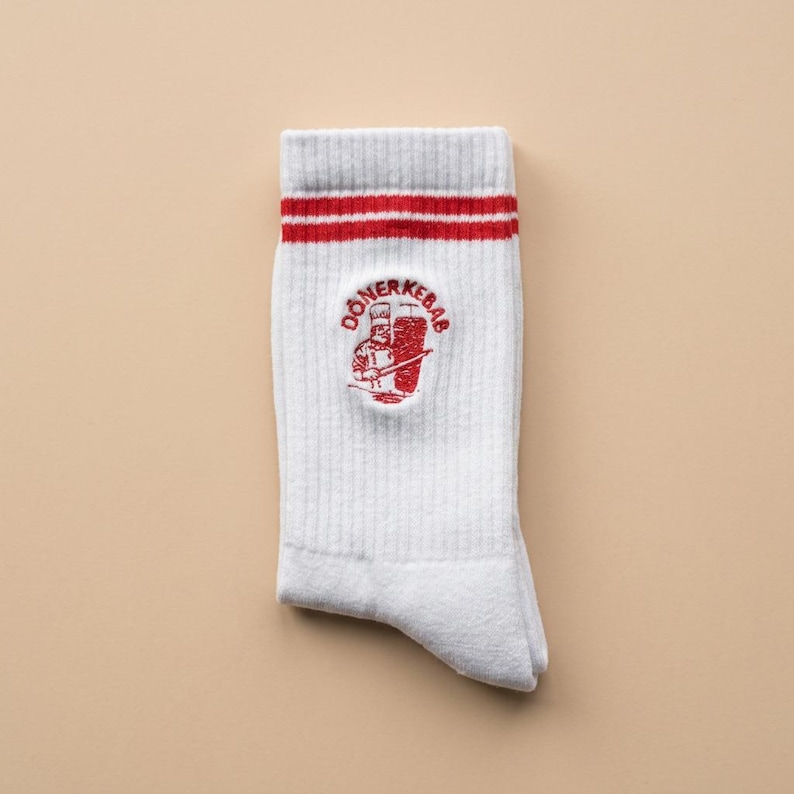 Pu&ograve; includere: Calzino sportivo bianco con strisce rosse nella parte superiore e un logo "DONER KEBAB" rosso ricamato. Il logo presenta uno chef e un kebab. Il calzino &egrave; realizzato in morbido materiale a coste.