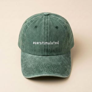 Puede incluir: Gorra de b&eacute;isbol verde con la palabra "overstimulated" bordada en blanco. La gorra tiene una visera curva y un dise&ntilde;o cl&aacute;sico de seis paneles. La tela tiene un aspecto ligeramente descolorido y vintage.