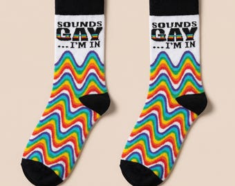 May include: Pair of white socks with black heels and toes. The socks feature the text "SOUNDS GAY... I'M IN" above a wavy rainbow pattern. The socks are designed with a colorful, eye-catching aesthetic.