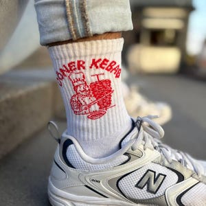 Könnte beinhalten: Weiße Socken mit dem roten Schriftzug "DONER KEBAB" und einer roten Grafik eines Kochs und eines Kebabs. Die Socken werden mit weißen und grauen Turnschuhen und hellblauen Jeans getragen.