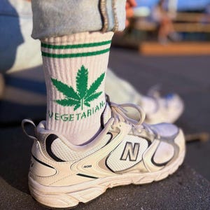 Op de afbeelding: Witte sportschoenen met zwarte en zilveren accenten, gecombineerd met witte sokken met een groen cannabisblad en het woord "VEGETARIAN". De schoenen hebben de letter "N" aan de zijkant.