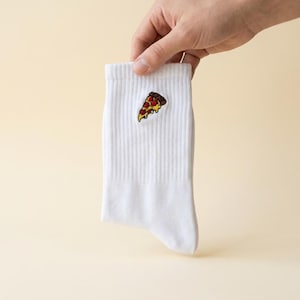 Pode incluir: Uma meia branca canelada com um pequeno gráfico bordado de uma fatia de pizza. A fatia de pizza tem uma crosta castanha, queijo derretido amarelo e coberturas de pepperoni vermelho. A meia é feita de tecido branco com textura canelada.