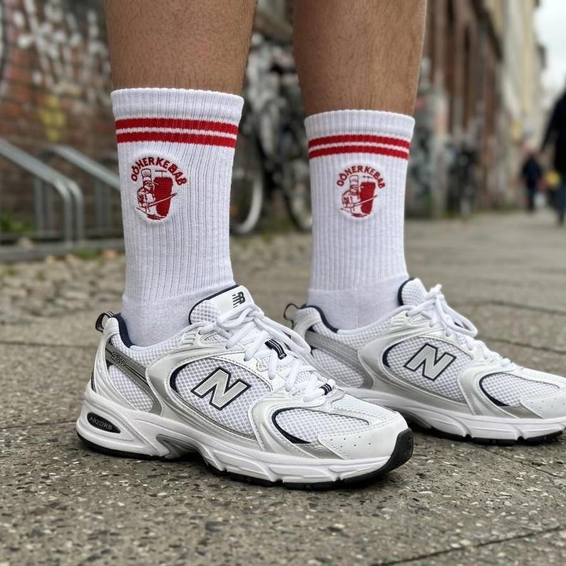 Pu&ograve; includere: Scarpe sportive bianche con dettagli argentati e suole nere, abbinate a calzini bianchi con strisce rosse e un logo. Le scarpe hanno la lettera "N" sul lato.