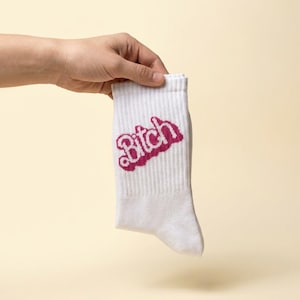 Könnte beinhalten: Eine weiße Socke mit Rippenstruktur und dem Wort Bitch, das in einer kräftigen, dunkelrosa Schreibschrift auf dem oberen Teil aufgedruckt ist. Die Socke hat eine Standardlänge und ein schlichtes Design mit einem klaren, gut lesbaren grafischen Element.