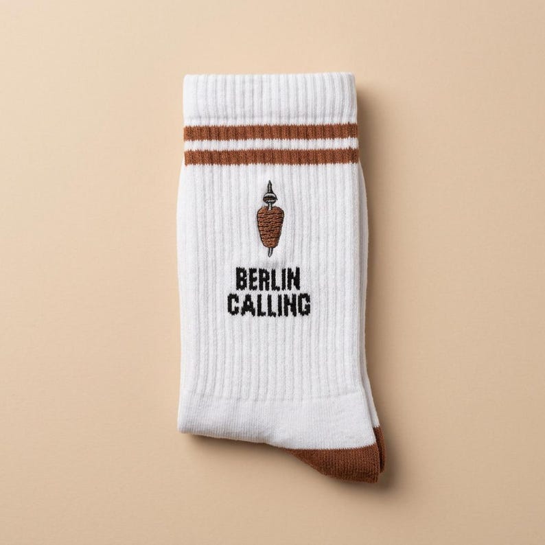 Pu&ograve; includere: Calzini bianchi con dettagli marroni, con la scritta "BERLIN CALLING" e un'immagine ricamata di un kebab. I calzini hanno strisce marroni nella parte superiore e sezioni di tallone e punta marroni. Realizzati in materiale a coste.