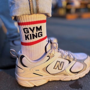 May include: White athletic socks with a red stripe and the text "GYM KING" in black. The socks are paired with white and silver athletic shoes. The shoes have the letter "N" on the side.