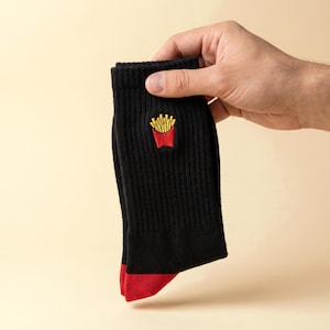Op de afbeelding: Een paar zwarte geribbelde enkelsokken met een kleine geborduurde afbeelding van gele frietjes in een rood bakje op het bovenste gedeelte. De sokken hebben een effen rood teengedeelte en een geribbelde textuur over het hele hoofdgedeelte.