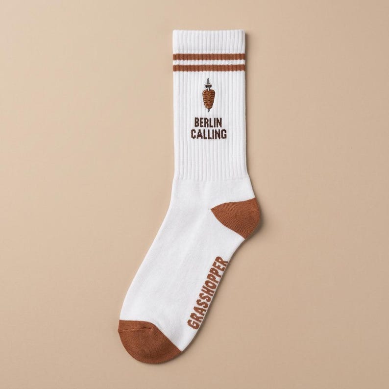 Pu&ograve; includere: Calzini bianchi con dettagli e strisce marroni. I calzini presentano la scritta "BERLIN CALLING" e una grafica di kebab. La parola "GRASSHOPPER" &egrave; stampata sul lato. Un accessorio divertente e unico.