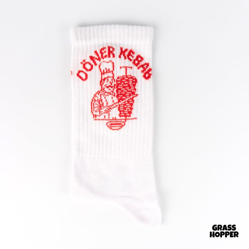 May include: White socks featuring a red graphic of a chef and a vertical meat skewer, with the words "DONER KEBAB" in red above the graphic. The socks are made of a soft material and are suitable for everyday wear.
