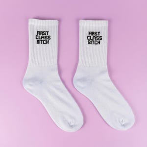 Könnte beinhalten: Zwei weiße, gerippte Crew-Socken mit dem schwarzen Aufdruck "FIRST CLASS BITCH". Die Socken sind vor einem hellvioletten Hintergrund platziert.