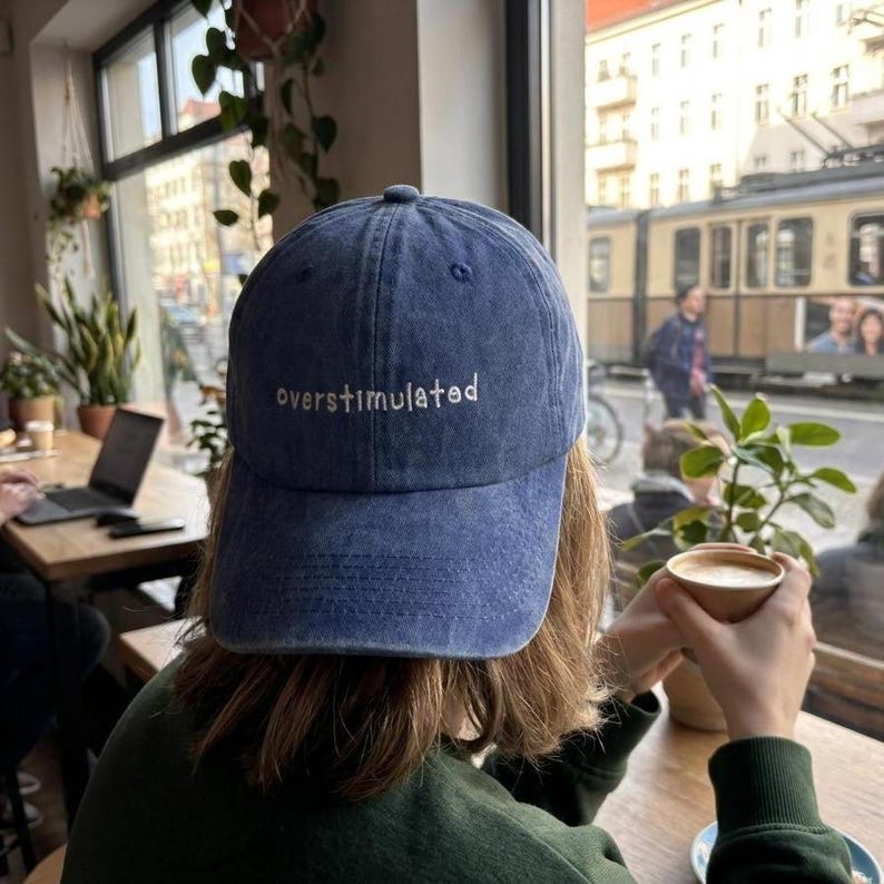 Puede incluir: Gorra de b&eacute;isbol de mezclilla azul con la palabra "overstimulated" bordada en blanco. La gorra la lleva una persona sentada a una mesa, sosteniendo una taza de caf&eacute;. El fondo muestra una ventana con una escena callejera.
